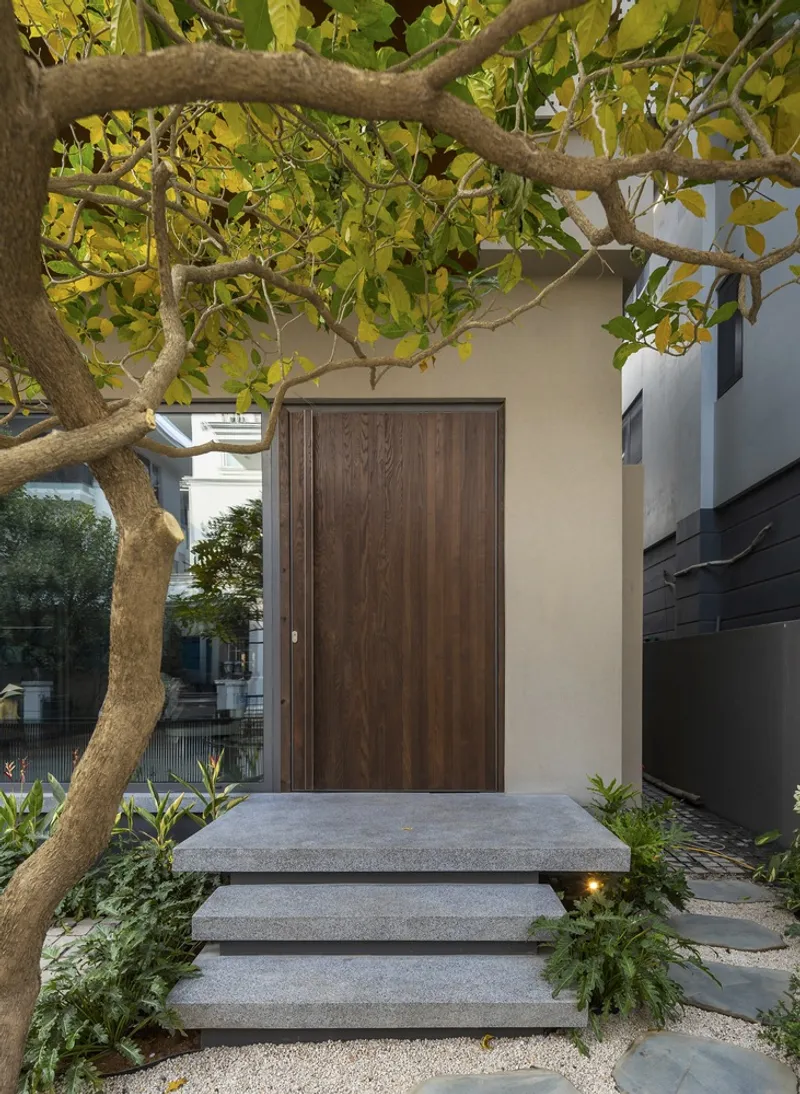 Timber entry door beneath overhanging tree branches with concrete steps and gravel paving below