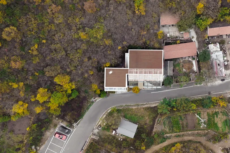 Aerial view of the timber-clad rooftop volumes beside a winding road and autumn forest canopy