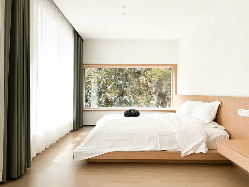 Bedroom with timber-framed horizontal window overlooking leafy trees and sheer grey curtains along the adjacent glazing