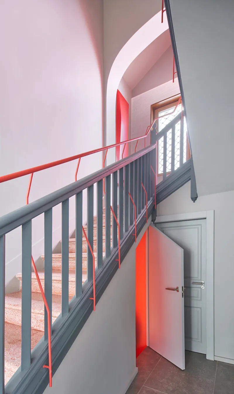 Helical staircase with grey metal balustrade and red handrail winding past pink-lit walls and doorways