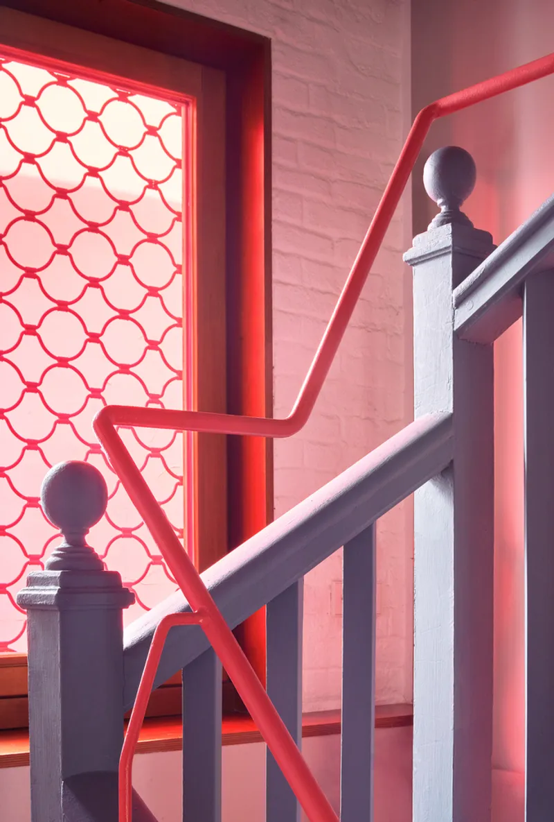 Staircase corner with red tubular handrail crossing grey balustrades beside a honeycomb-patterned screen in red light