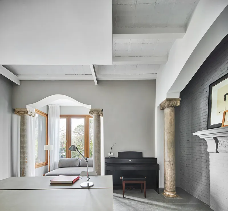 Open living space with ionic columns flanking an arched window and upright piano beneath concrete beams