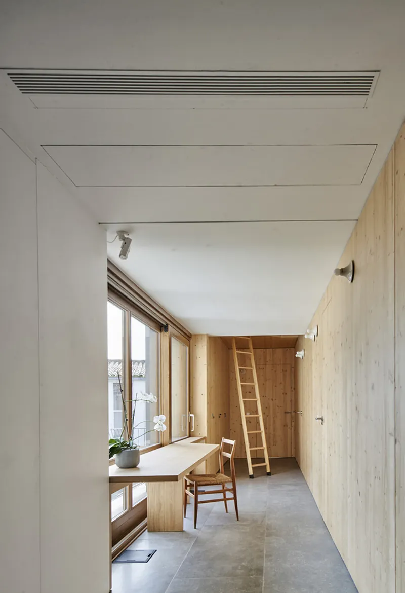 Narrow corridor with timber-lined walls and ladder leading to window desk beneath linear ventilation grille