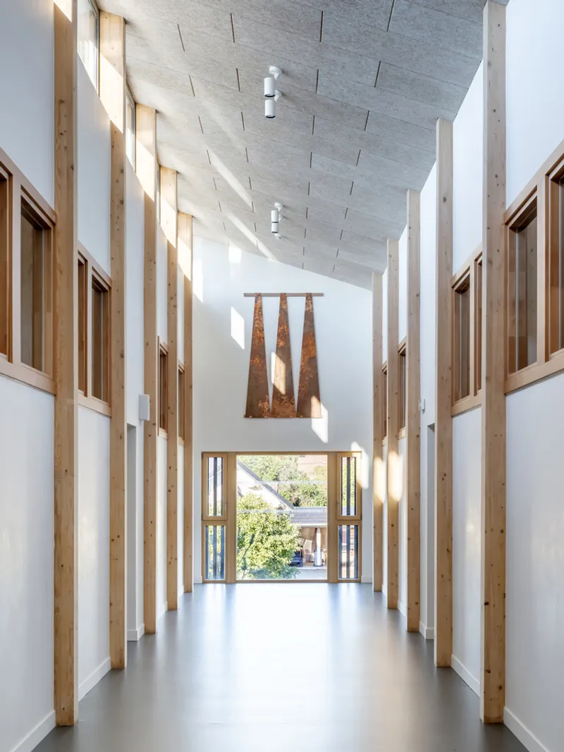 Double-height corridor with timber-framed walls and acoustic ceiling panels leading to glazed doors and courtyard beyond