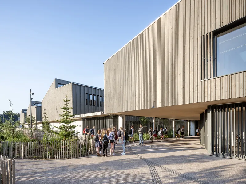 Cantilevered timber-clad volume above a covered walkway with students gathering in afternoon light