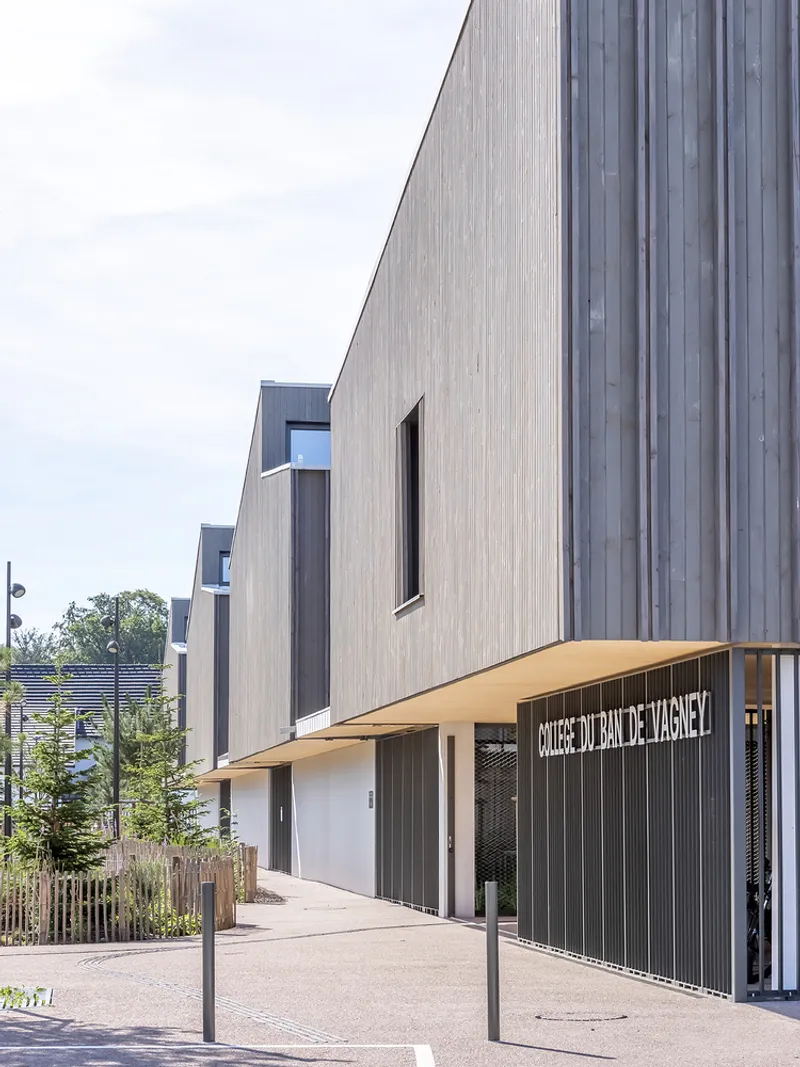 Timber-clad facade with overhanging canopy sheltering an entrance beside planted beds and grasses