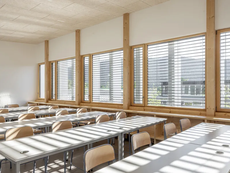 Classroom interior with timber columns and row of windows casting slatted shadows across tables