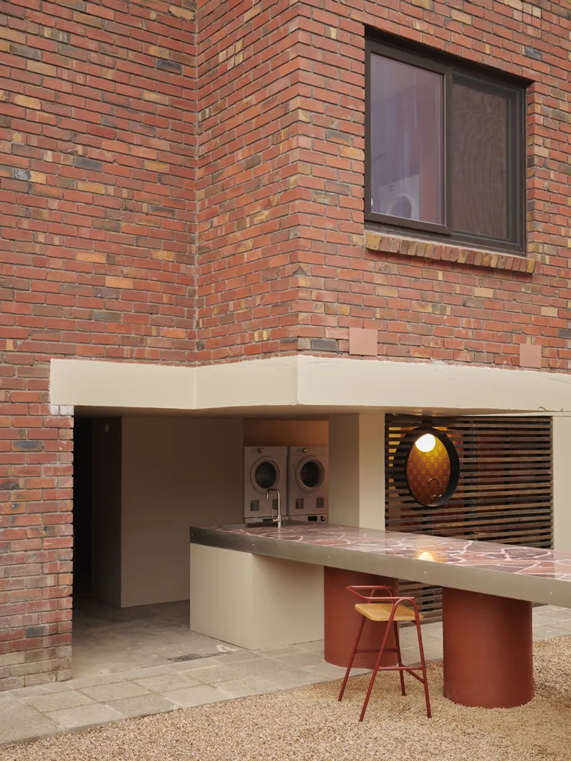 Ground-floor communal laundry niche with white bench counter and red cylindrical base beneath variegated brick wall