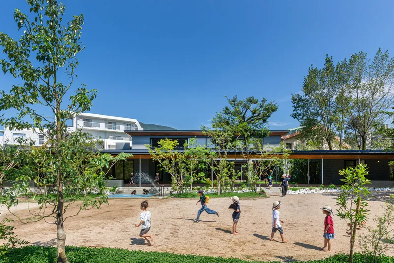 Sand play area surrounded by young trees with horizontal pavilion roofline under clear blue sky