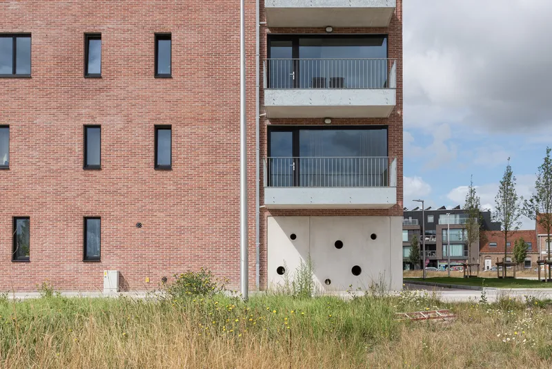 White concrete base with circular openings beneath stacked balconies and red brick facade in summer grasses