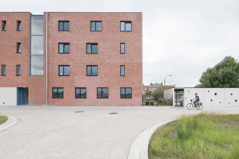Street view of brick facade with cyclist passing white perforated wall along curved paved plaza