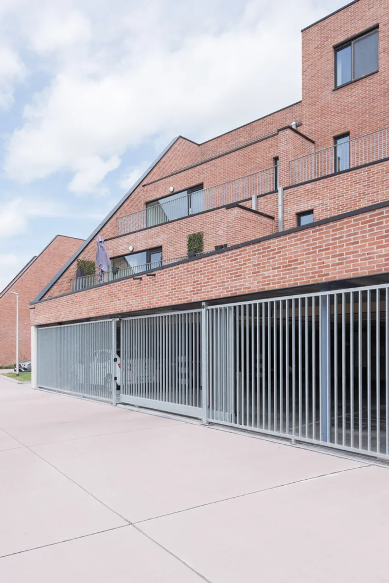 Stepped brick facade with inset balconies above a long vertical-slat metal fence under cloudy skies
