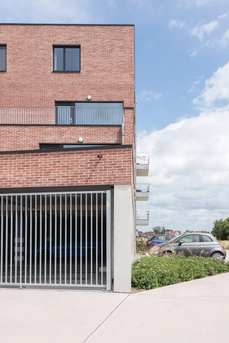 Brick volume with vertical-slat metal screen at base beneath stacked balconies and cloudy afternoon sky