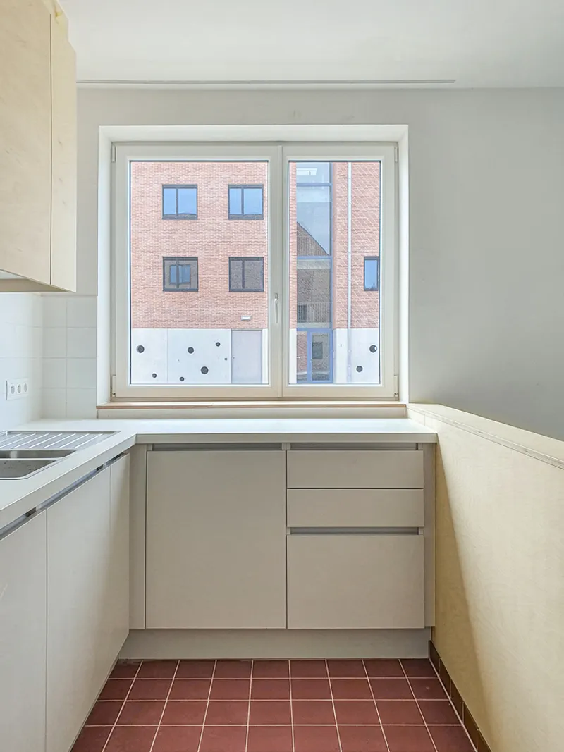 Kitchen window framing view of the brick courtyard facade with light wood cabinetry and terracotta floor