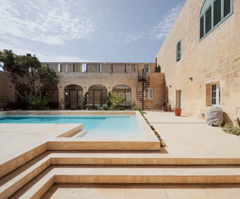 Courtyard pool with limestone steps and arched colonnade beyond under scattered clouds