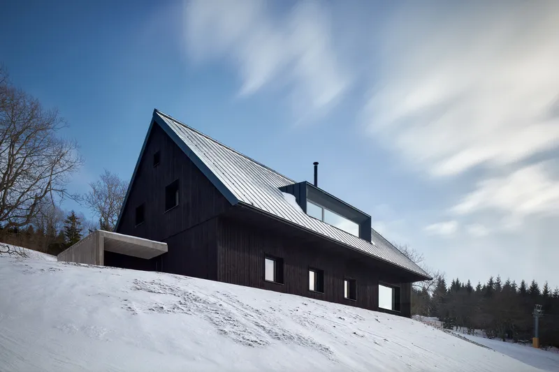 Black timber-clad gabled volume with vertical cladding and metal roof on a snowy hillside