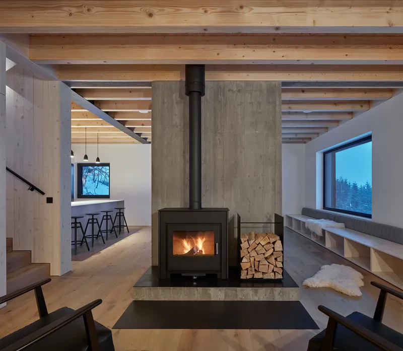Central wood-burning stove with concrete surround and stacked firewood beneath exposed timber ceiling beams