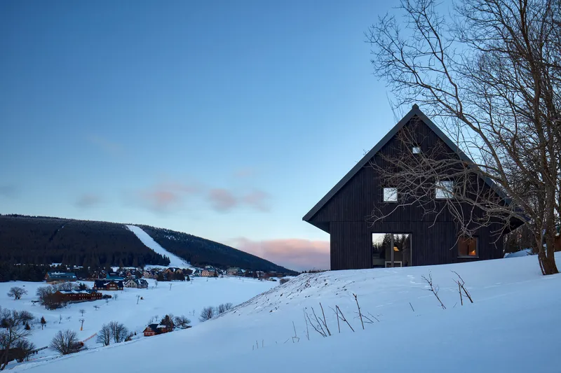 Dark timber-clad gable volume on a snow-covered hillside at dusk with bare trees