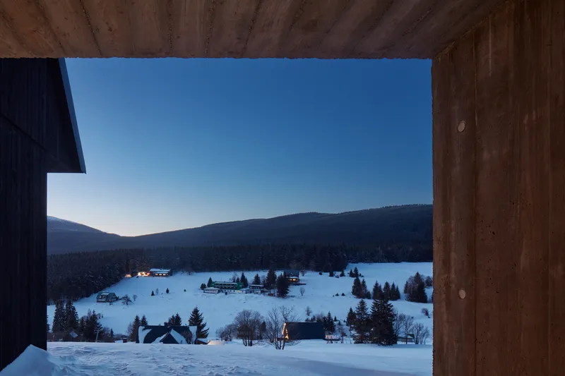 Framed view through timber soffit opening toward a snowy valley with scattered buildings at dusk