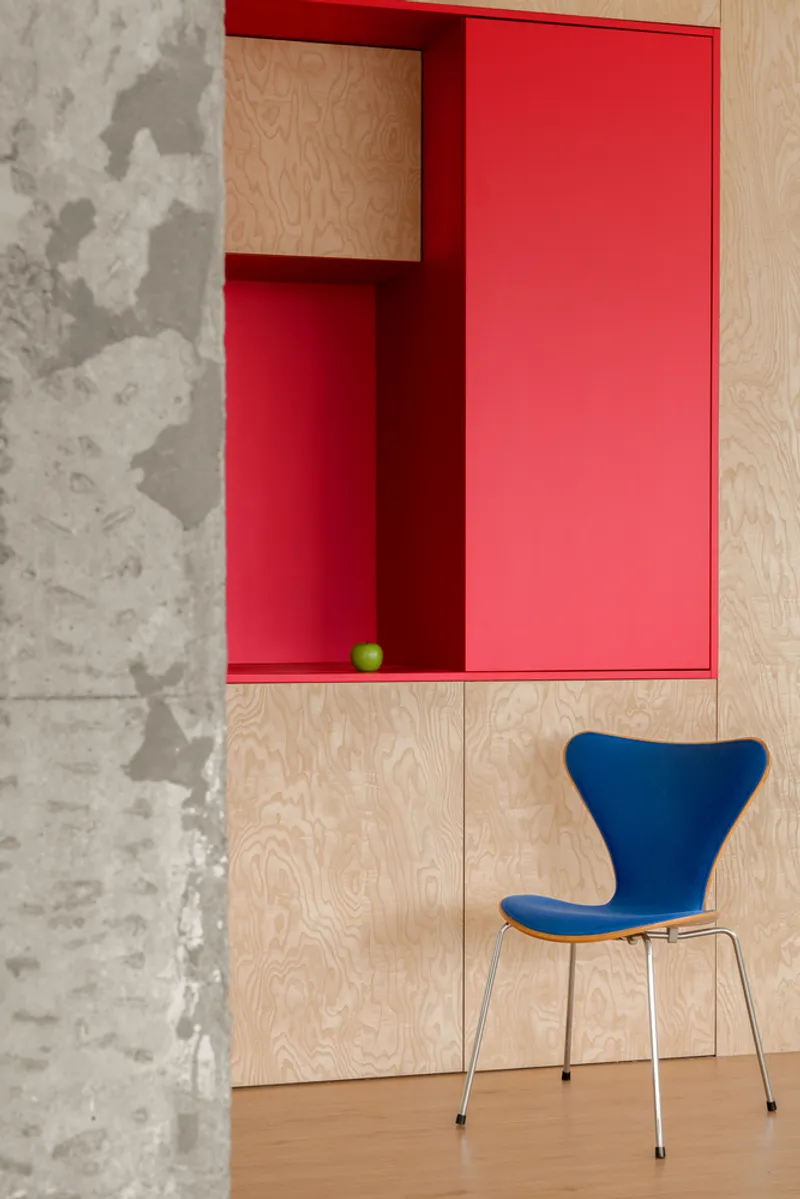Blue upholstered chair beside a plywood wall with red inset shelving and a green apple