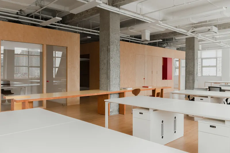 Workstation area with white desks and plywood partitions beneath exposed mechanical ducts and concrete beams