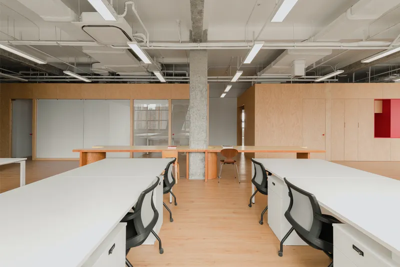 Open office space with white desks, exposed concrete columns and plywood partition walls with translucent panels