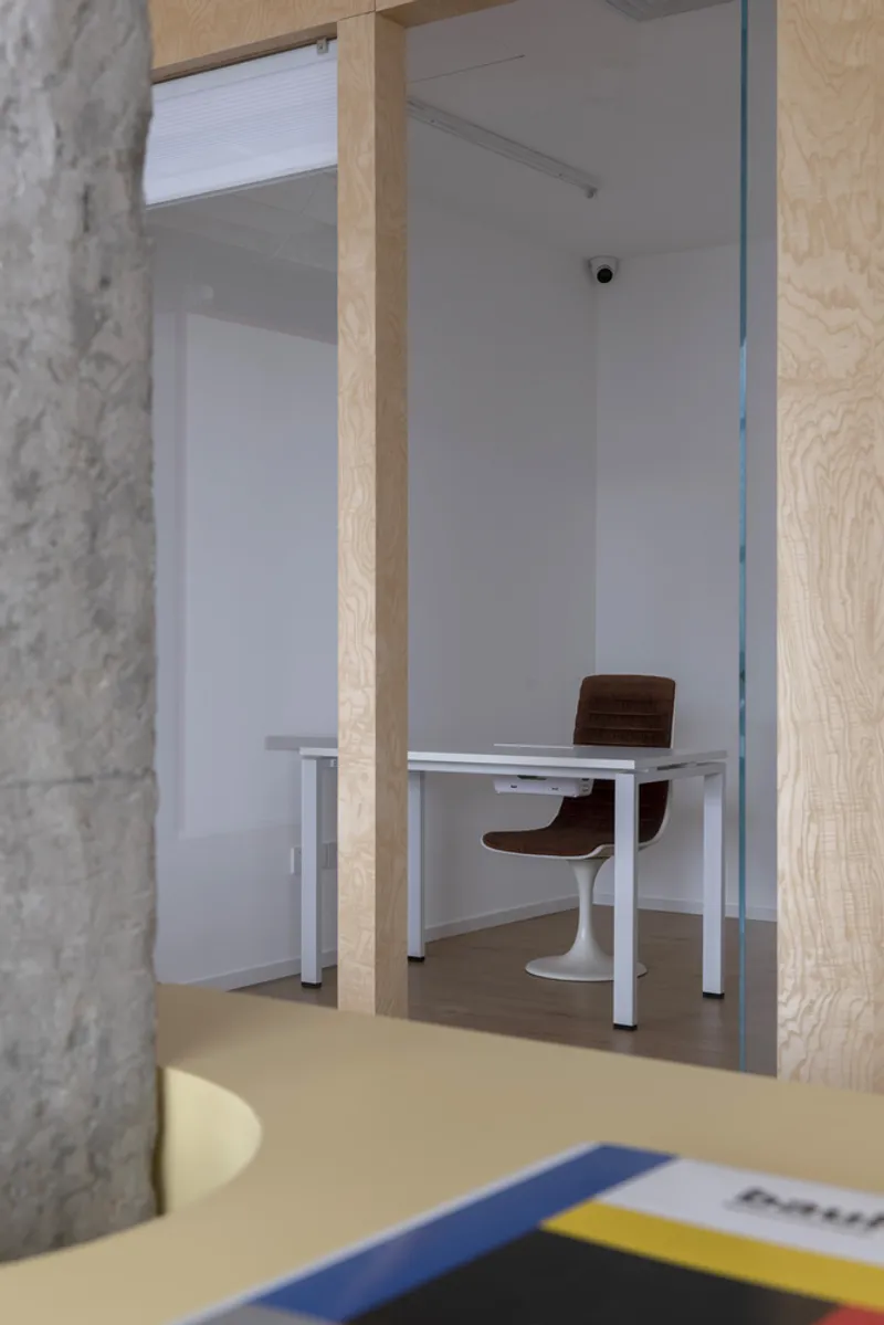 View through plywood framed opening into a small office room with white desk and brown chairs