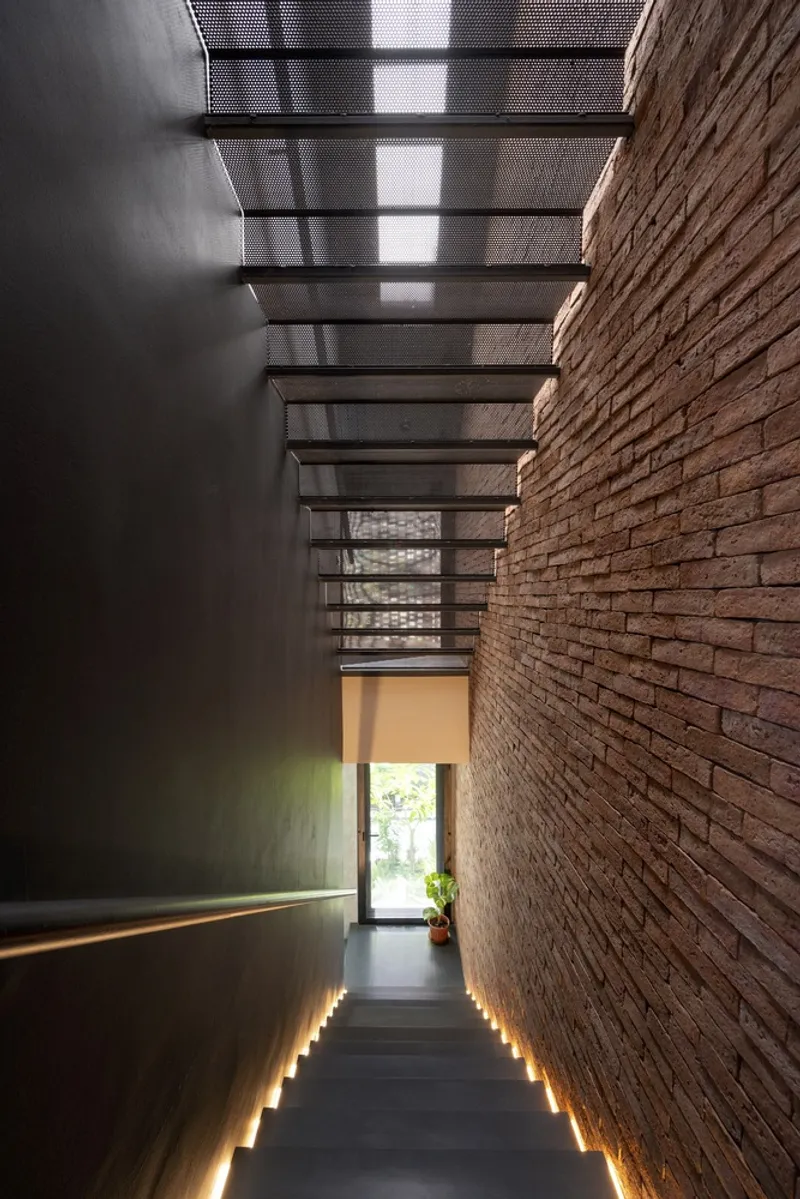 Descending staircase with lit treads between dark wall panel and brick wall under mesh skylight
