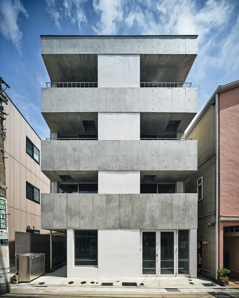 Street facade of a four-story building with stacked concrete slabs and recessed white balconies under blue sky