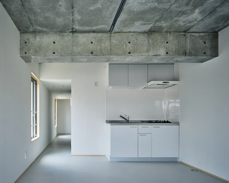 Interior view of compact kitchen unit below exposed concrete beam with visible formwork ties and ceiling soffit