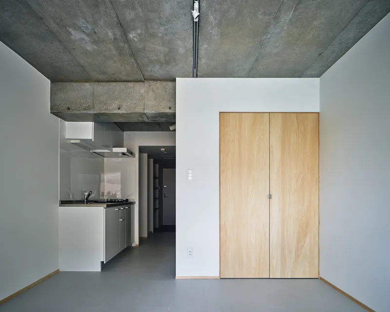 Living space with light wood closet doors and kitchen alcove beneath raw concrete ceiling structure