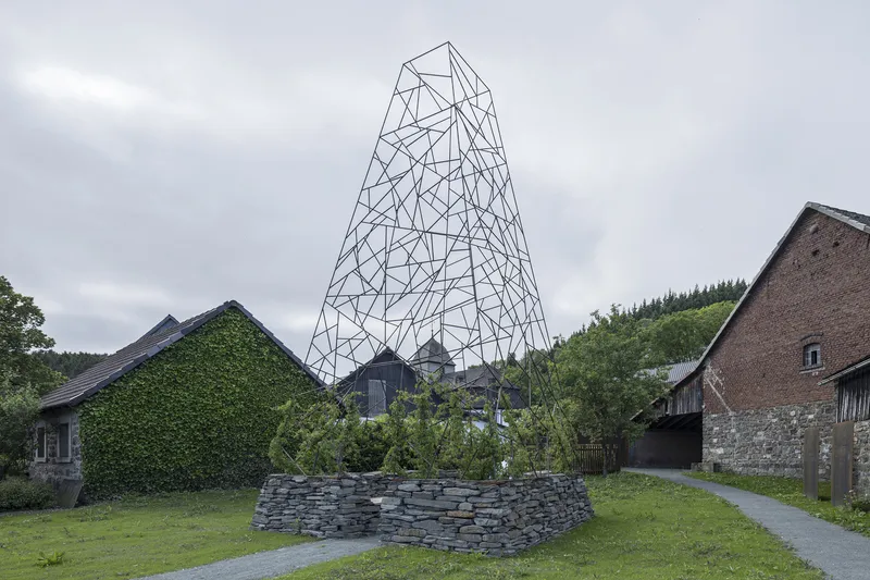 Tapered steel wireframe tower rising above a layered slate stone base and ivy-covered farm buildings
