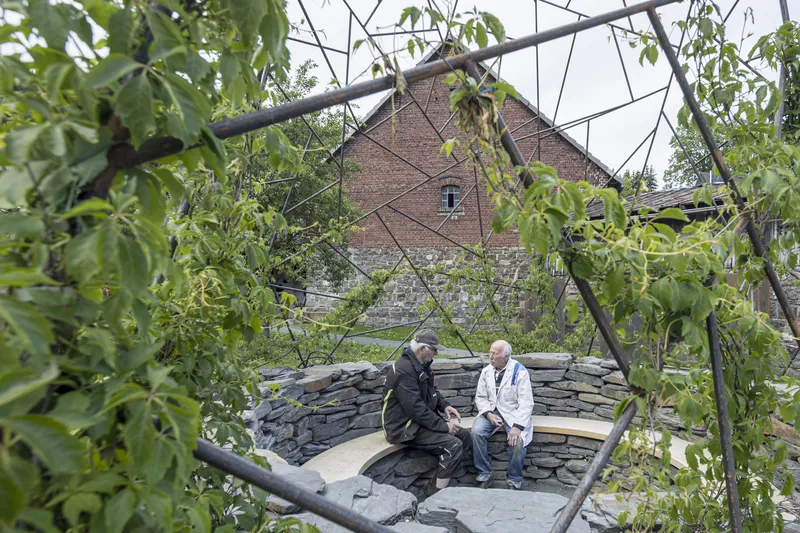 Two visitors seated in the slate-walled enclosure beneath the triangulated steel lattice and fruit tree branches