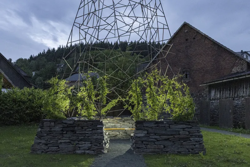 Geometric steel framework sheltering young trees planted in the layered stone enclosure at dusk