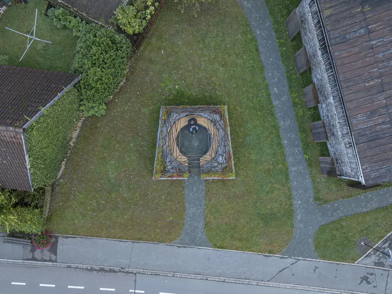 Drone view showing the square stone enclosure in a grass lawn between paved paths and timber barns