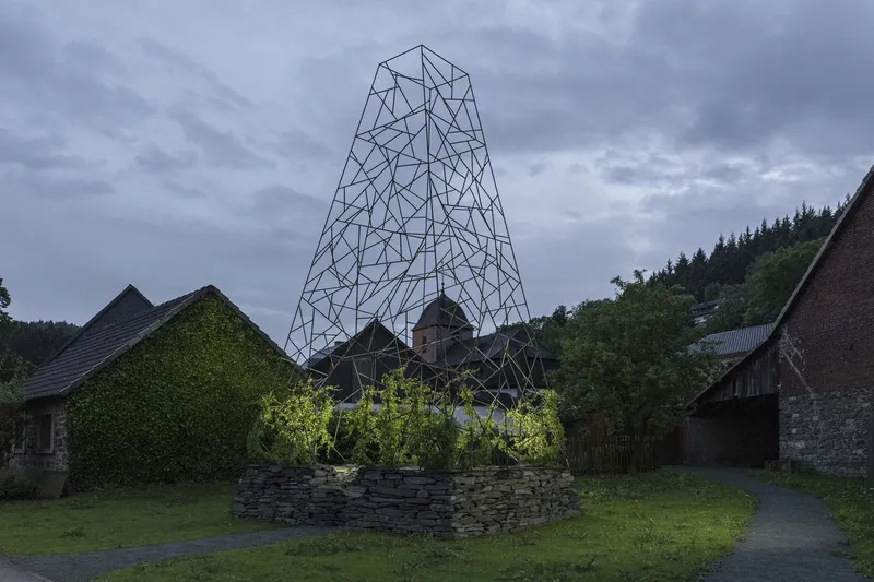 The wireframe tower and stone planting bed under an overcast evening sky beside rural farm buildings