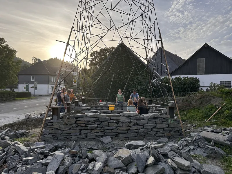 Dry-stone foundation walls with triangulated metal frame tower at dusk with community members working
