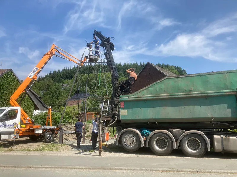 Orange crane lifting a steel framework structure above a green dump truck near forested hills