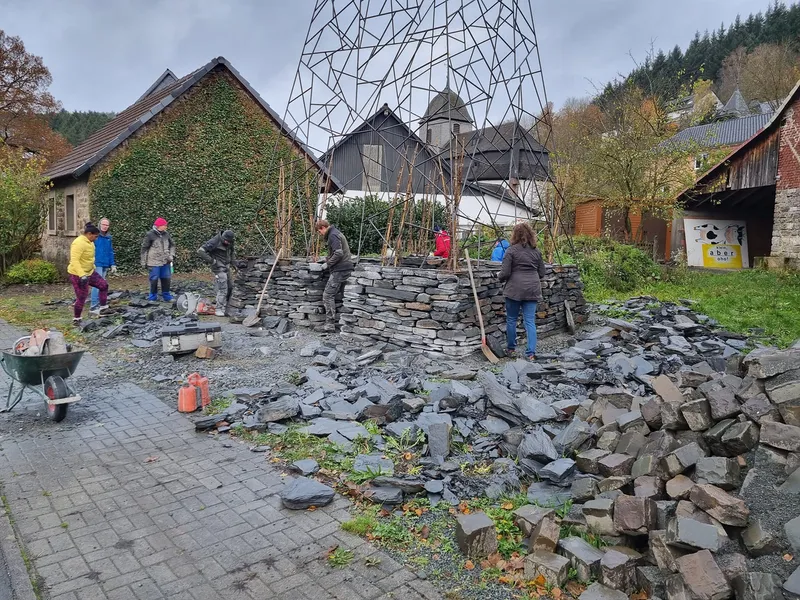 Group constructing a dry-stacked slate stone wall around a pyramidal steel tower on an overcast day
