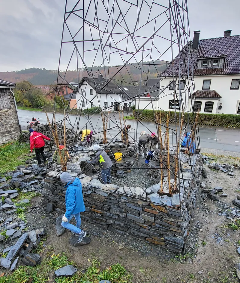 Circular dry-stone wall base with triangulated metal frame tower under construction, workers in rain gear
