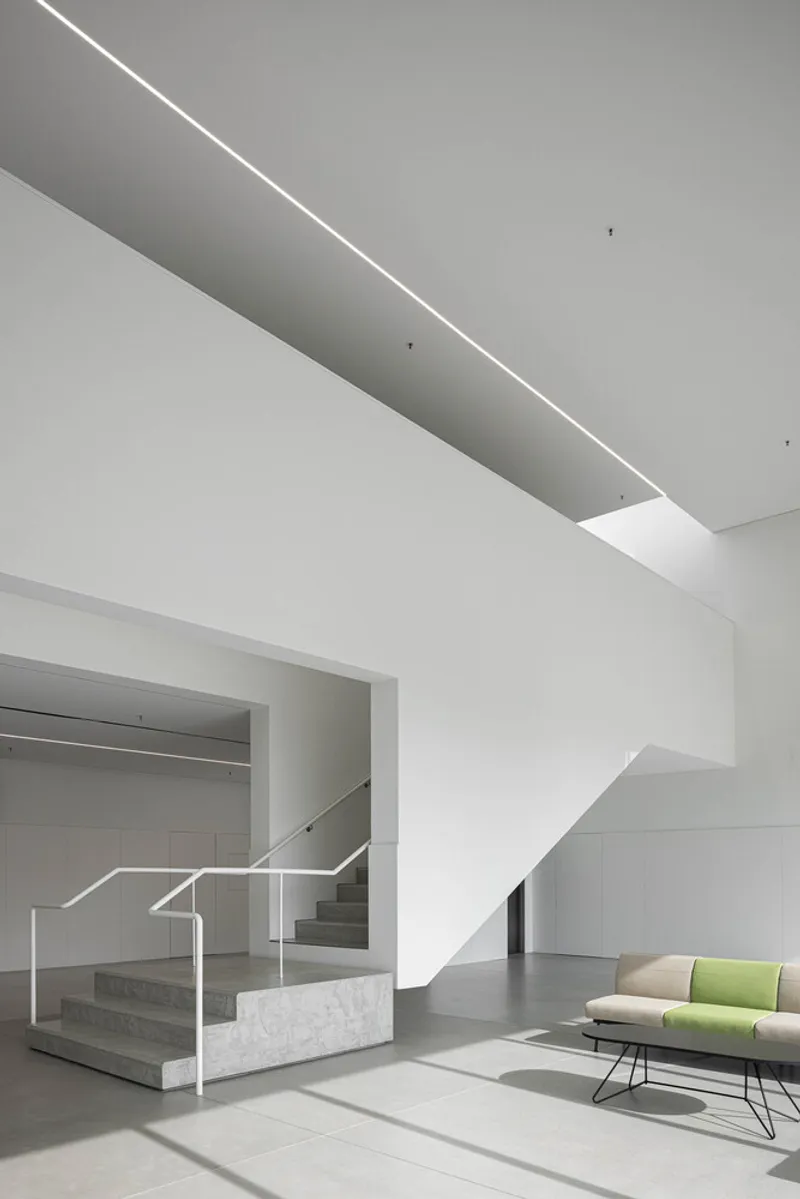 Interior staircase with marble treads and white metal railing beneath a linear skylight casting diagonal shadows