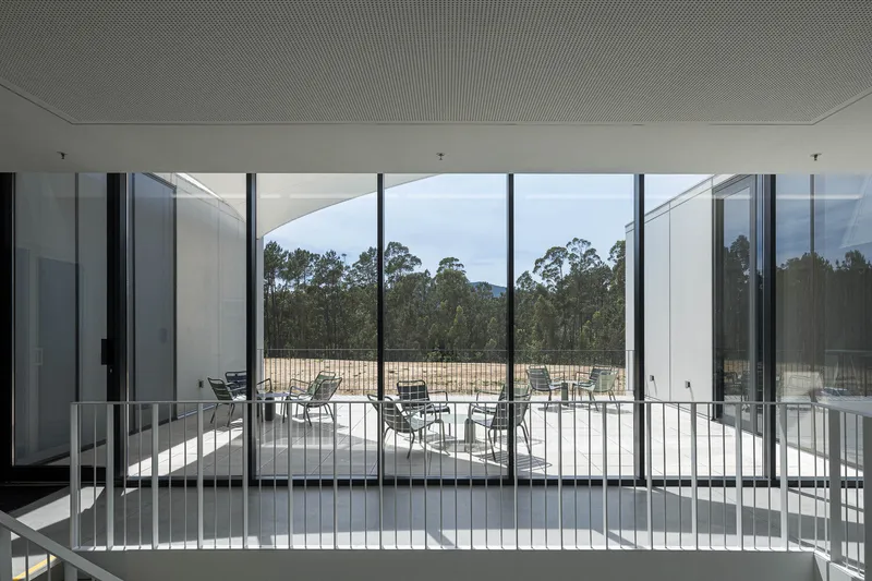 Interior balcony with white metal railing overlooking outdoor terrace and tree line through floor-to-ceiling glass