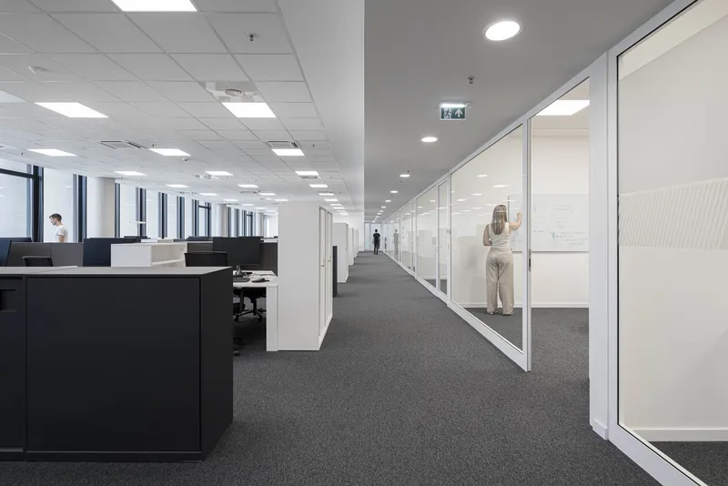 Office corridor with glass-walled meeting rooms and workstations beneath suspended ceiling panels and recessed lighting
