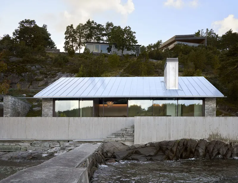 Waterside facade with standing seam metal roof and stacked stone piers behind a concrete retaining wall