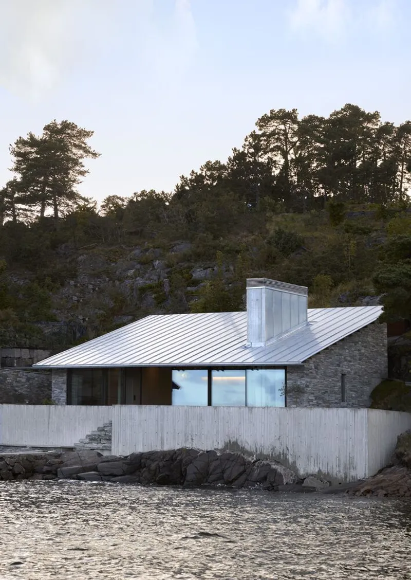 Lakeside elevation at dusk showing the metal roof and central chimney mass against the rocky hillside