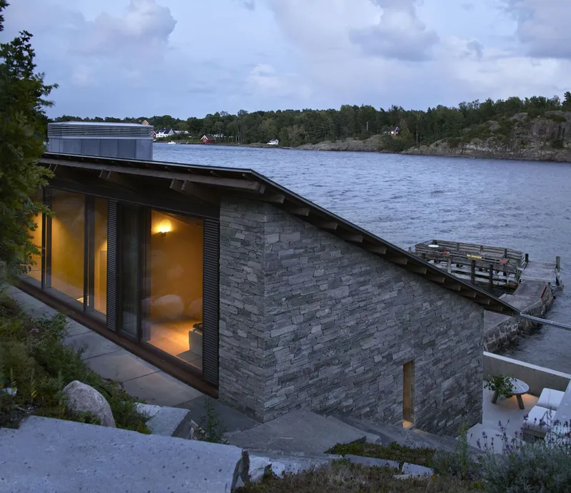 Stone-clad volume with sloped metal roof and timber dock at dusk on the water