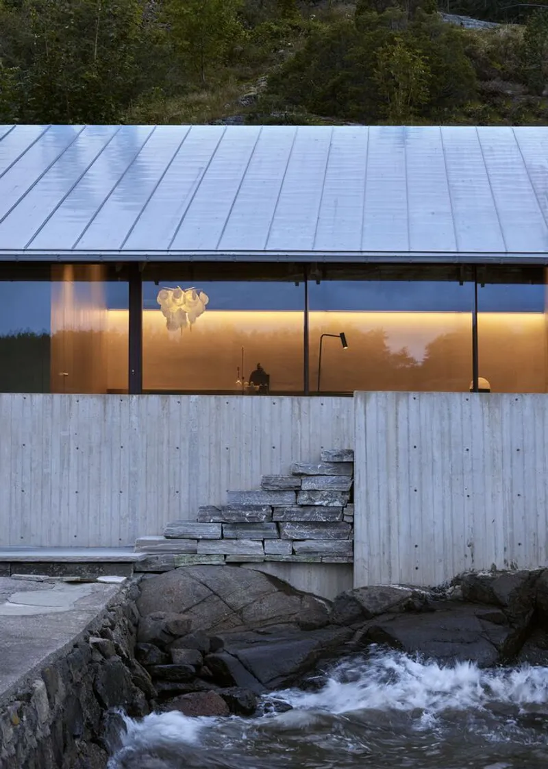 Glazed facade with board-formed concrete base and stone steps at water's edge at dusk