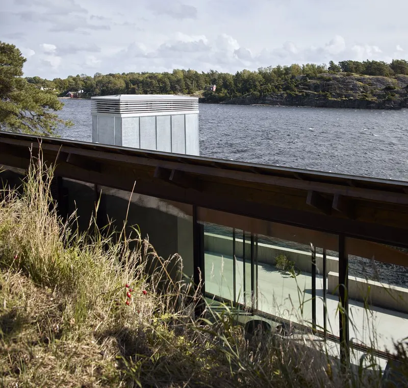 Low-angled view of the glass-walled volume with flat roof nestled into rocky terrain beside the water
