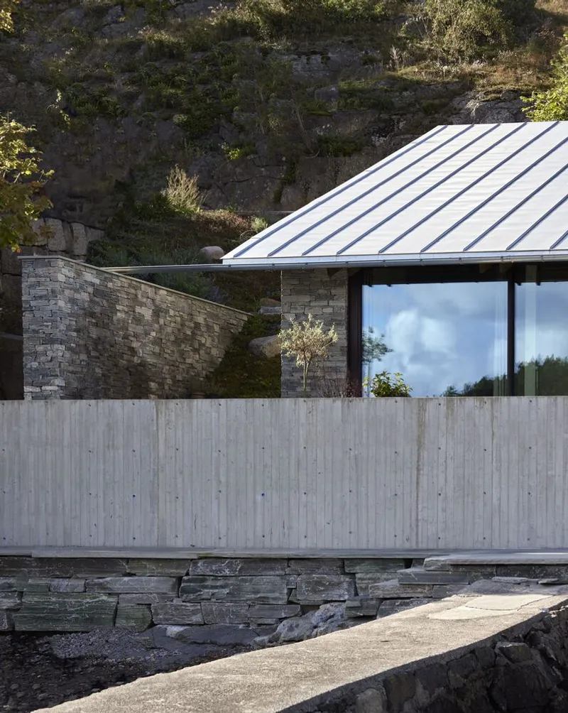 Board-formed concrete wall and stacked stone base beneath a sloped metal roof against hillside rock