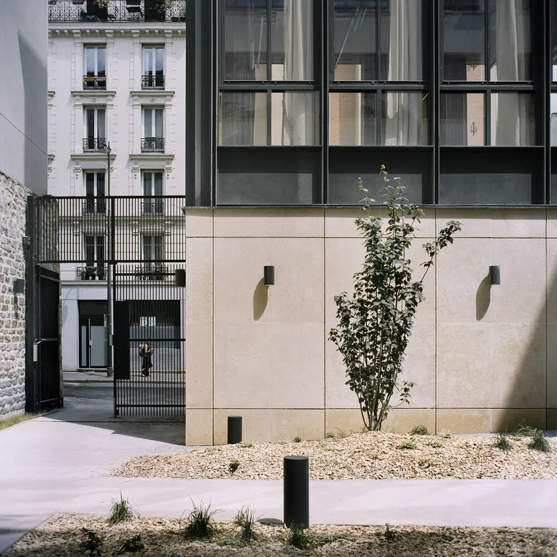 Street view of the beige stone facade with planted gravel bed and a young tree beside the entry passage